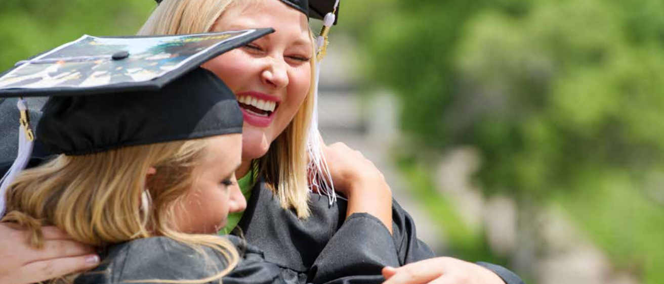Two female graduates wearing mortarboards, smiling and hugging.