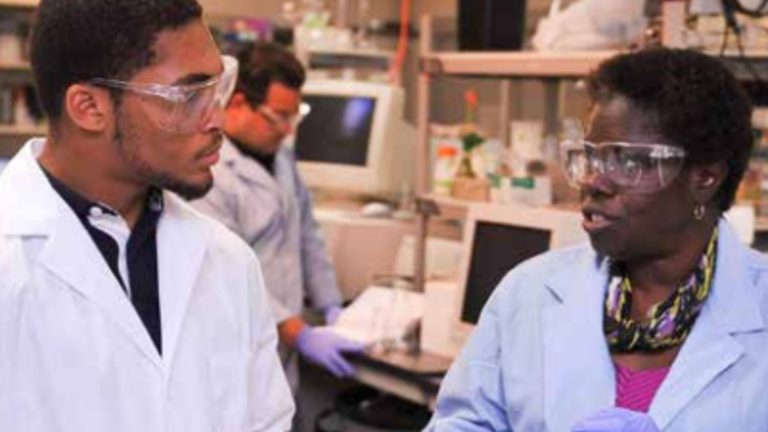 Two researchers in lab coats and safety glasses, talking.
